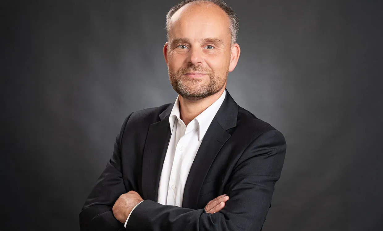 Business-Porträt eines älteren Mannes mit verschränkten Armen, weißem Hemd und schwarzem Sakko vor anthrazitfarbenem Hintergrund in Schleswig, mit ernstem Blick und leichtem Schmunzeln.